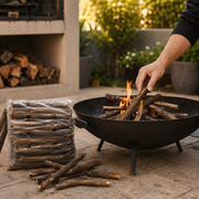 Person adding firewood to a pit fire, surrounded by stacked logs and greenery.
